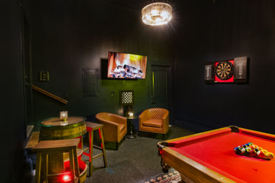 Cozy basement game room with a red-felt pool table, wall-mounted dartboard, two tan leather armchairs, barrel-style pub table with stools, TV showing a fireplace, and a sparkling chandelier against dark walls.
