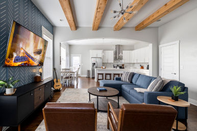 Modern open-concept living room and kitchen with exposed wooden ceiling beams, blue sectional sofa, leather armchairs, wall-mounted TV showing a roaring fire, round wood coffee table, hardwood floors, kitchen island with stools, white cabinets and nearby dining area with a guitar.