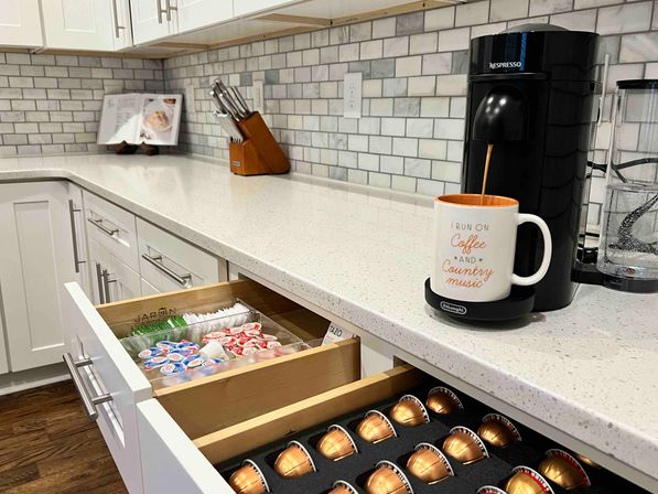 Modern white kitchen coffee station: black single-serve pod machine pouring into a mug labeled 'I run on coffee and country music', open drawers with organized metallic coffee capsules and creamer cups on a white quartz countertop and subway-tile backsplash.