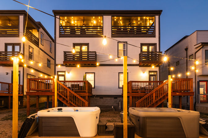 Cozy dusk scene of modern three-story residential townhouses with illuminated string lights, wooden decks and balconies, and two hot tubs on the backyard patio
