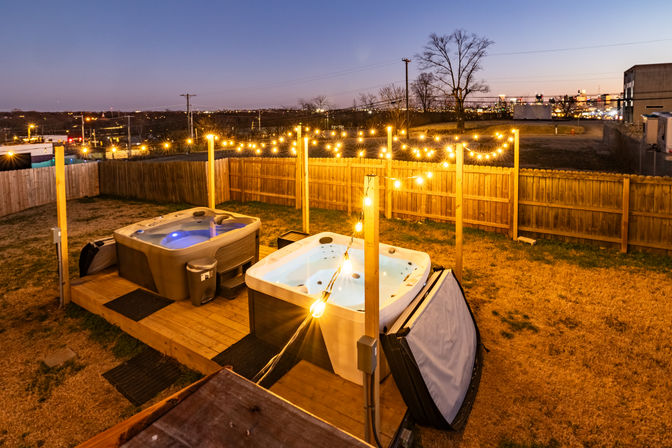 Two soaking hot tubs on a raised wooden deck in a fenced backyard, glowing string lights overhead and a distant city skyline at dusk.