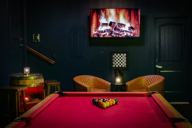 Cozy game room interior with red-felt pool table racked for a break, wall-mounted TV showing a fireplace, two leather lounge chairs, barrel side table and wall chessboard against dark walls.