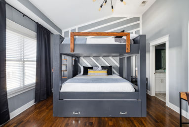 Bright modern bedroom featuring a built-in dark gray bunk bed with wooden top rail, diagonal striped accent wall, hardwood floors, black curtains, decorative pillows and an open doorway to a bathroom.