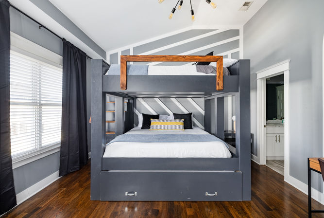 Bright modern bedroom featuring a built-in dark gray bunk bed with wooden top rail, diagonal striped accent wall, hardwood floors, black curtains, decorative pillows and an open doorway to a bathroom.
