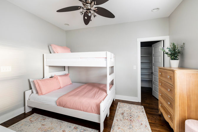 Bright modern bedroom with white twin-over-full bunk bed dressed in pink bedding, hardwood floors, patterned rugs, wooden dresser with potted plant, and dark-blade ceiling fan