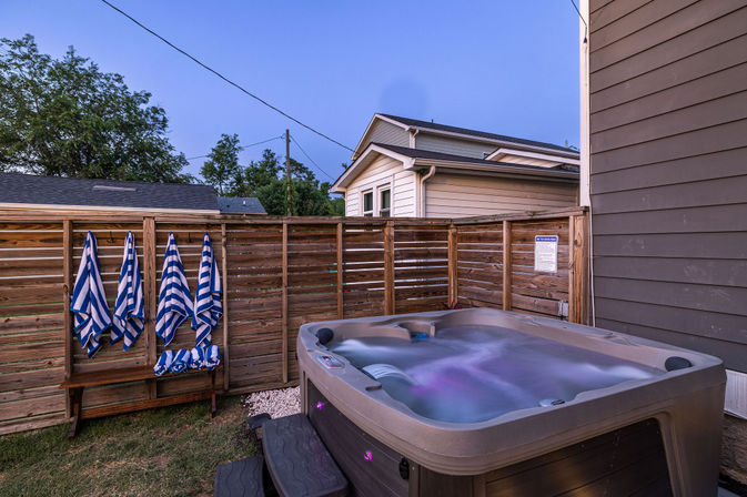 Twilight suburban backyard featuring a bubbling hot tub lit with purple LEDs, a wooden privacy fence, bench with blue-and-white striped towels, and house siding in the background.