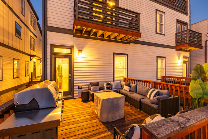 Inviting urban apartment balcony at dusk with wooden deck, L-shaped outdoor sectional and cushions, covered coffee table, gas grill, potted plants and warm wall lighting.