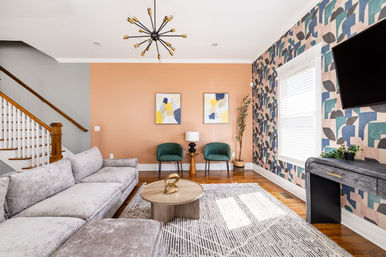 Bright modern living room with peach accent wall and playful geometric wallpaper, light-gray sectional sofa, round wooden coffee table with sculptural decor, two green accent chairs, mid-century starburst chandelier, hardwood floors and a sunlit window.