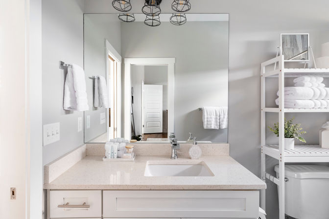 Bright, airy gray-and-white residential bathroom with quartz vanity and undermount sink, large mirror, cage pendant lights, wall towels and open shelving stacked with white towels and a potted plant.