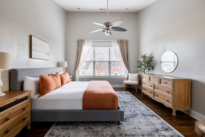 Bright contemporary master bedroom with large window and sheer curtains, gray upholstered bed with rust-orange pillows and runner, wooden nightstands and dresser, ceiling fan, patterned area rug, hardwood floors and a potted plant.
