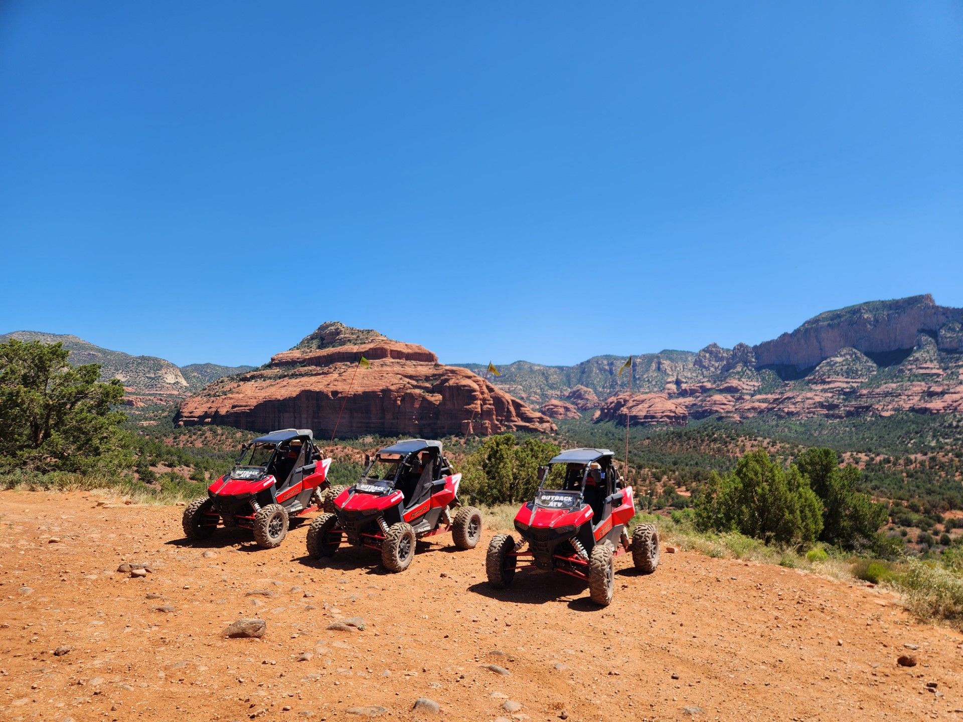 Three red off‑road UTVs parked on a sunlit red‑dirt overlook with layered red‑rock mesas, scattered junipers and a vast clear blue sky — ready for a desert off‑road adventure.