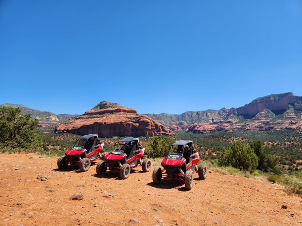 Three red off‑road UTVs parked on a sunlit red‑dirt overlook with layered red‑rock mesas, scattered junipers and a vast clear blue sky — ready for a desert off‑road adventure.