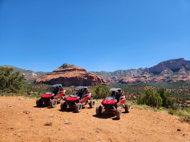 Three red off‑road UTVs parked on a sunlit red‑dirt overlook with layered red‑rock mesas, scattered junipers and a vast clear blue sky — ready for a desert off‑road adventure.