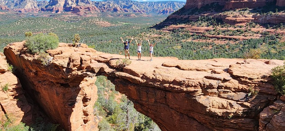 Three hikers with arms raised standing on a sandstone natural arch overlooking Sedona, Arizona red-rock canyon and green forest under a clear blue sky