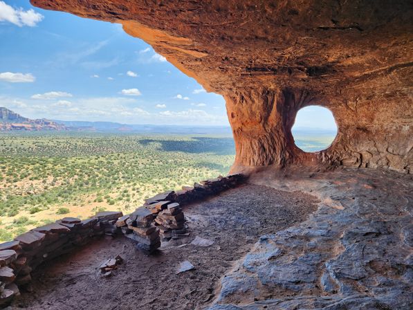 View from a red sandstone cave with a circular natural window peeking out over a vast Southwestern desert valley, scattered scrub, and distant red-rock mesas.