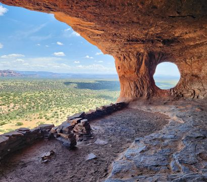 View from a red sandstone cave with a circular natural window peeking out over a vast Southwestern desert valley, scattered scrub, and distant red-rock mesas.