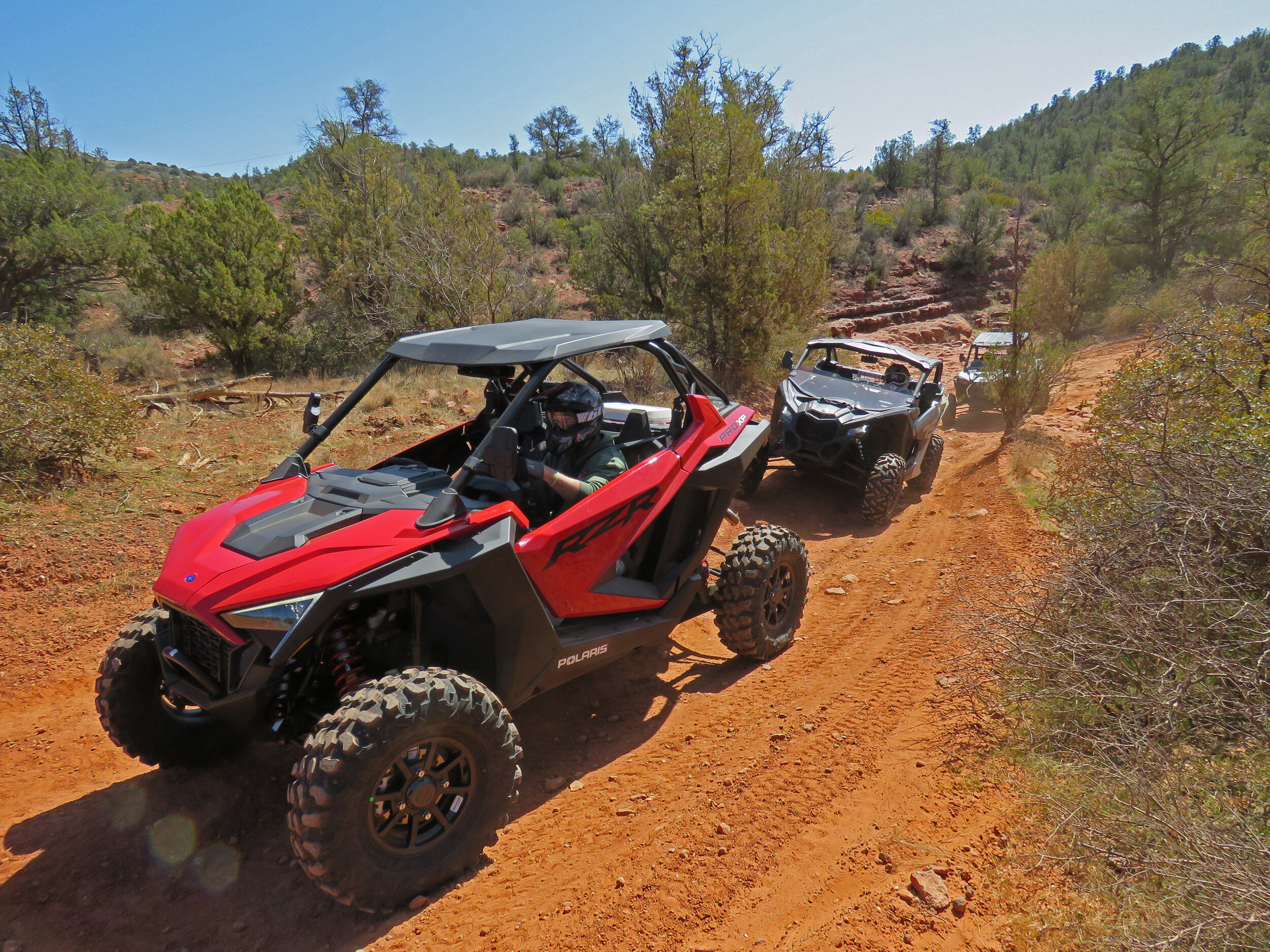 Red side-by-side UTV leading two more on a dusty red-dirt off-road trail through Southwestern red-rock desert scrub and low canyon hills under a clear blue sky.