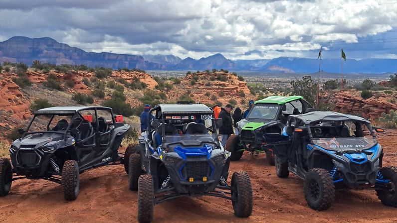 Several off-road UTVs parked on a red-rock desert overlook with people, rugged mesas in the distance and a dramatic cloudy sky