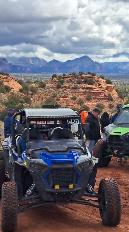 Several off-road UTVs parked on a red-rock desert overlook with people, rugged mesas in the distance and a dramatic cloudy sky