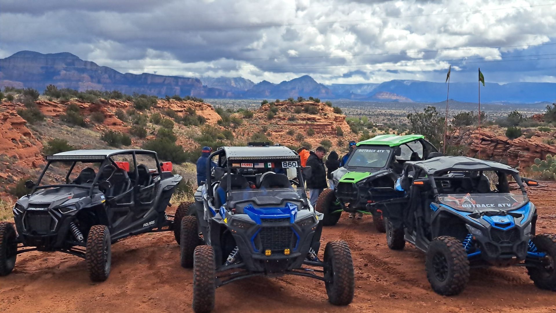 Several off-road UTVs parked on a red-rock desert overlook with people, rugged mesas in the distance and a dramatic cloudy sky