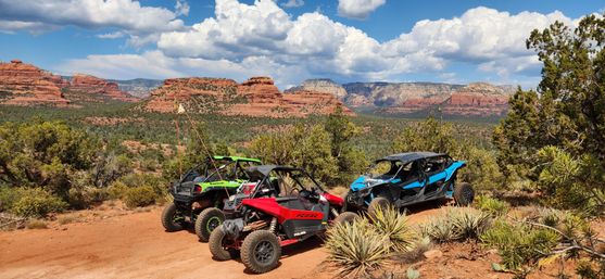 Three colorful off-road side-by-side UTVs parked on a dusty desert trail among yucca and juniper, overlooking Sedona, Arizona red-rock mesas under a blue sky with puffy clouds.