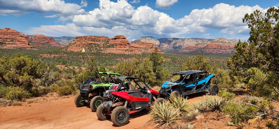 Three colorful off-road side-by-side UTVs parked on a dusty desert trail among yucca and juniper, overlooking Sedona, Arizona red-rock mesas under a blue sky with puffy clouds.