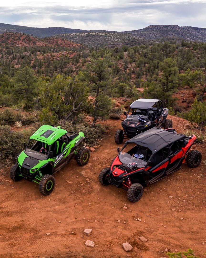 Three colorful off-road side-by-side UTVs (green, black, red) parked on a red-dirt trail amid juniper and scrub with red-rock mesas and wide open sky in a Southwestern desert landscape.