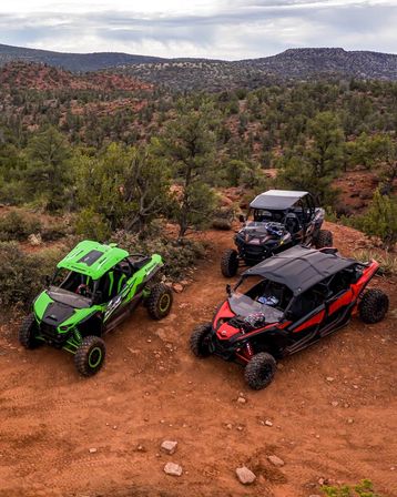 Three colorful off-road side-by-side UTVs (green, black, red) parked on a red-dirt trail amid juniper and scrub with red-rock mesas and wide open sky in a Southwestern desert landscape.