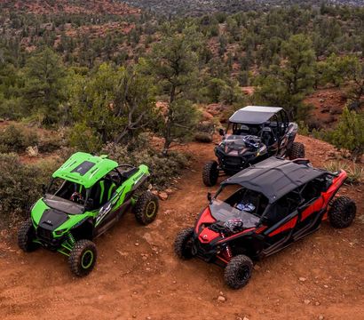 Three colorful off-road side-by-side UTVs (green, black, red) parked on a red-dirt trail amid juniper and scrub with red-rock mesas and wide open sky in a Southwestern desert landscape.