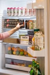 Open home kitchen refrigerator with a hand reaching for a canned drink among rows of canned beverages, egg cartons, takeout containers and berries, clear drawers of citrus and apples, and bottled drinks in the door.