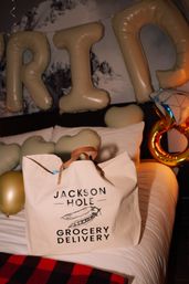 Cream canvas grocery delivery tote with leather handles on a bed, surrounded by gold letter balloons, heart-shaped cushions and a ring-shaped foil balloon in a festive hotel-room setting