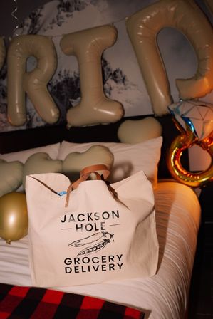 Cream canvas grocery delivery tote with leather handles on a bed, surrounded by gold letter balloons, heart-shaped cushions and a ring-shaped foil balloon in a festive hotel-room setting