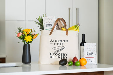Canvas grocery delivery tote with leather handles on a modern white kitchen island, filled with bananas, pineapple, coffee bag and bread, surrounded by a wine bottle, avocado, lime, orange, pear and a small vase of bright mixed flowers.