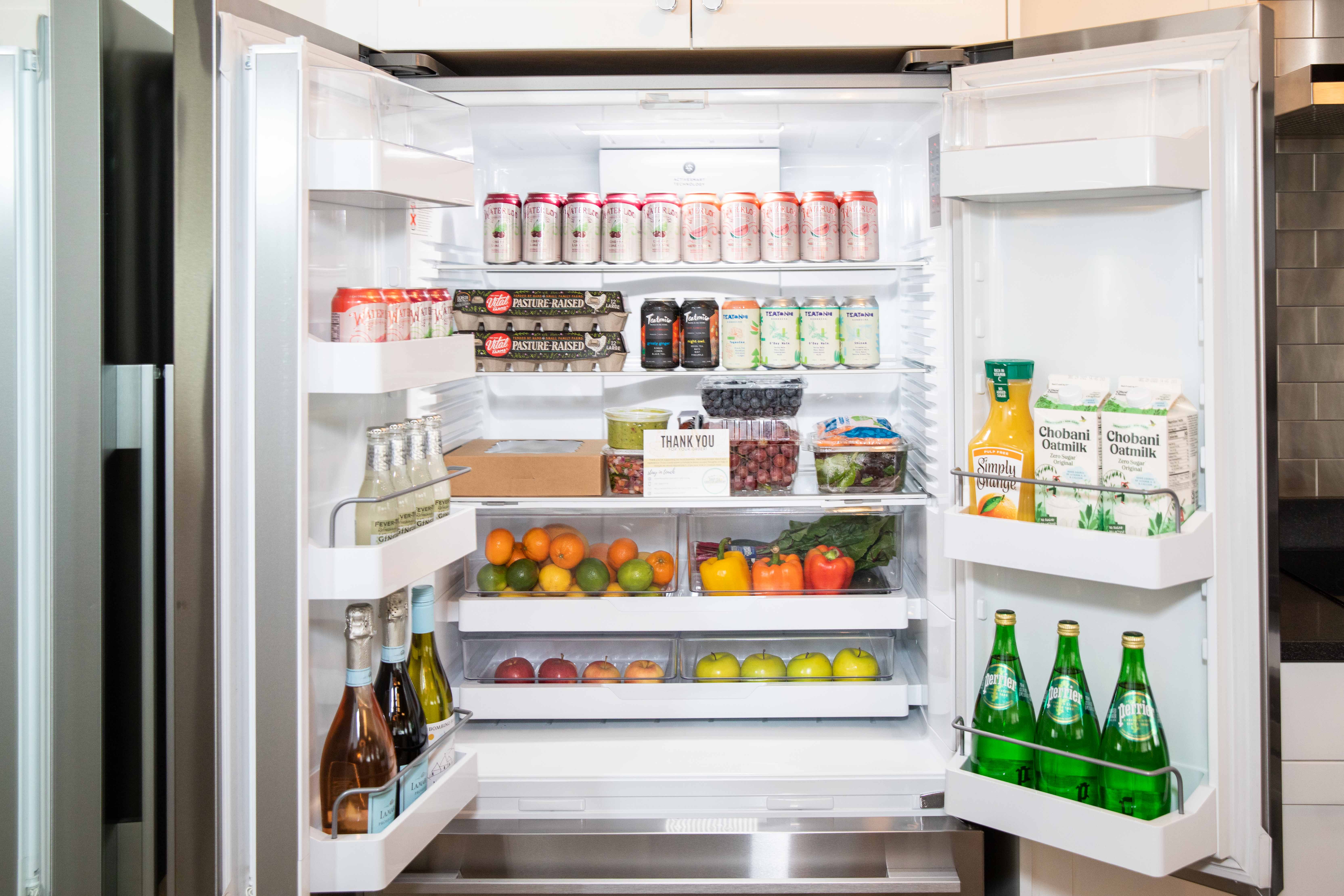 Open stainless-steel refrigerator in a modern kitchen, neatly stocked with canned drinks, sparkling water, wine, oat milk, orange juice, eggs, berries, apples, citrus and fresh vegetables.