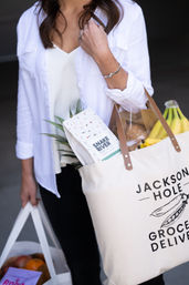 Casual shopper in a white shirt carrying a reusable canvas grocery tote and bag brimming with bananas, pineapple, fresh bread and a bag of coffee — eco-friendly grocery delivery vibe.
