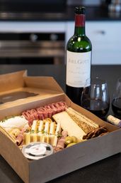 Takeout charcuterie box on a modern kitchen countertop with assorted cheeses, rolled cured meats, crackers, olives and nuts, paired with a bottle of red wine and two filled glasses.