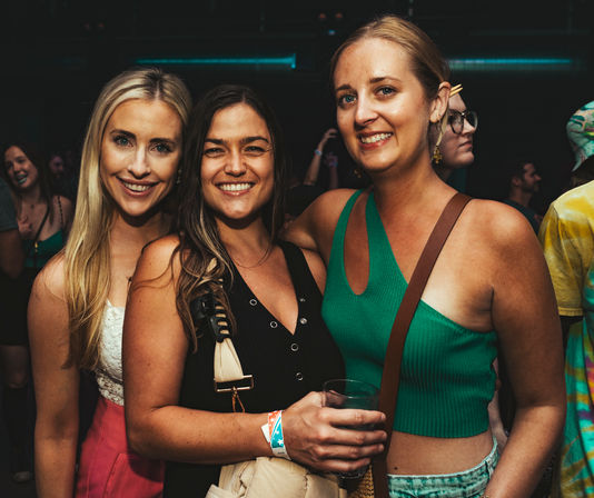 Three smiling women posing at a crowded indoor music venue during a night out, one holding a drink