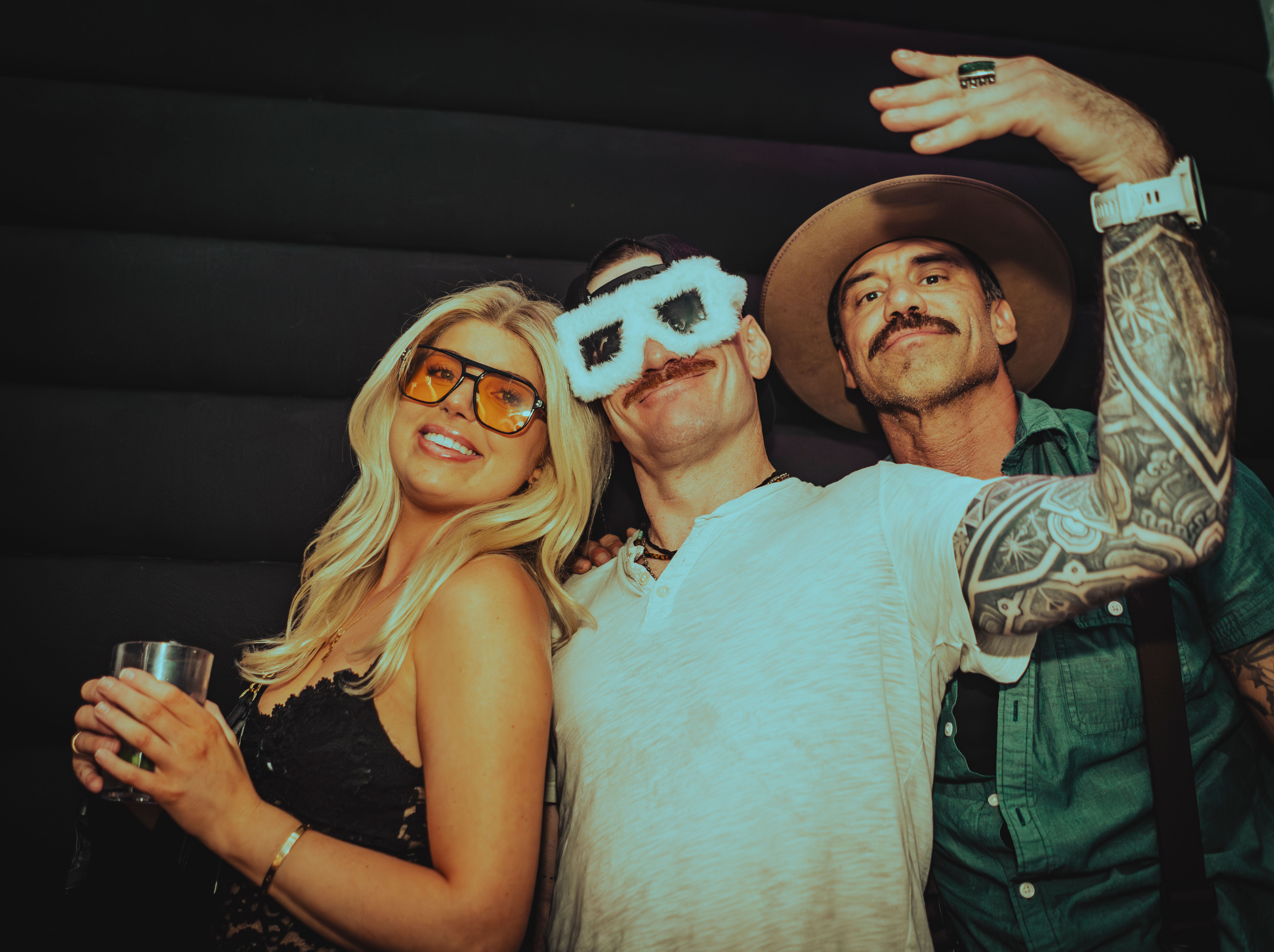 Three friends in a nightclub photo booth — blonde woman with yellow sunglasses holding a drink, man in fluffy novelty glasses, and a tattooed man wearing a wide-brim hat, fun nightlife vibe