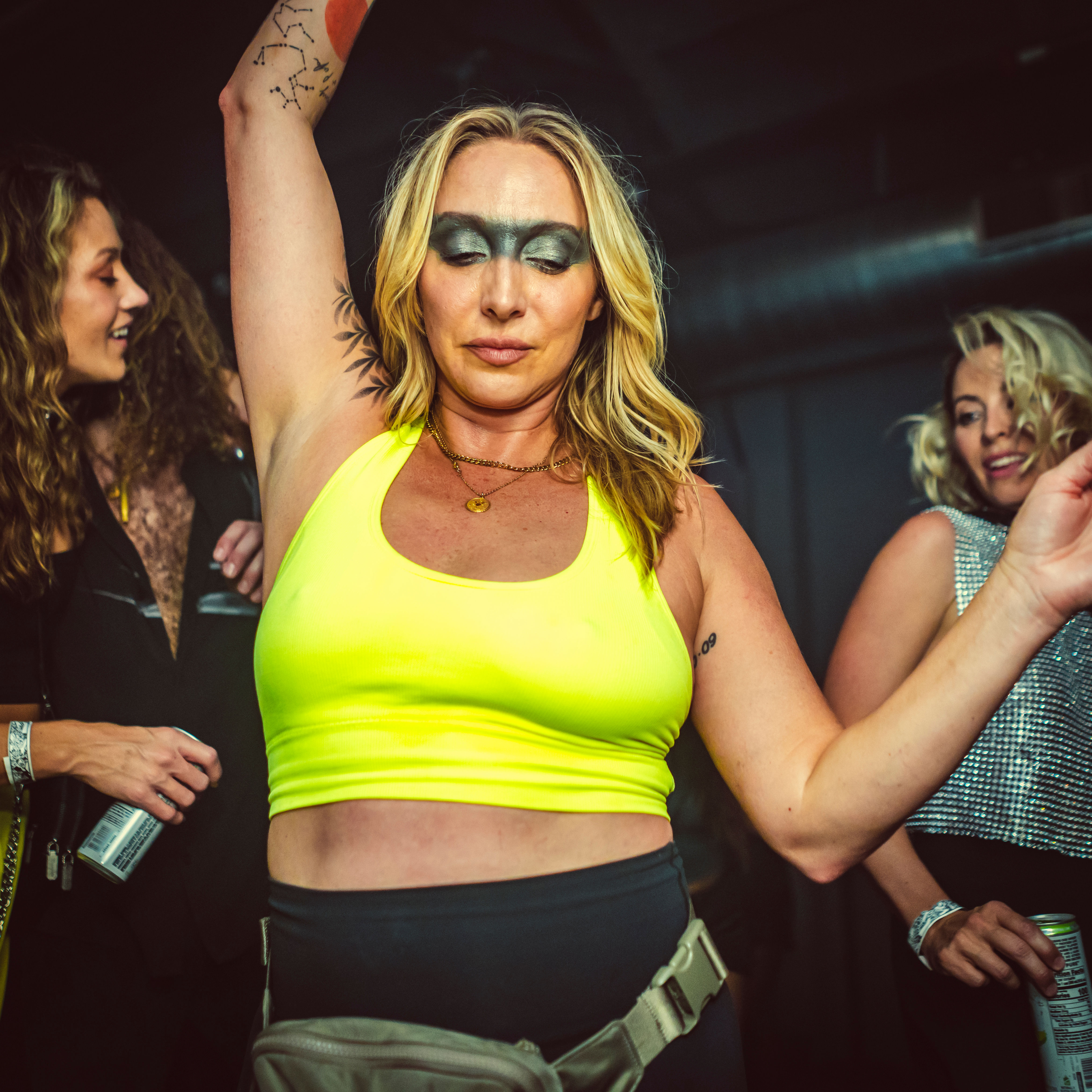 Woman in a neon yellow crop top and bold metallic eye makeup dancing with arm raised on a crowded indoor club dance floor, wearing a fanny pack and surrounded by friends with canned drinks
