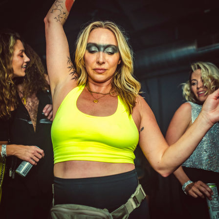 Woman in a neon yellow crop top and bold metallic eye makeup dancing with arm raised on a crowded indoor club dance floor, wearing a fanny pack and surrounded by friends with canned drinks