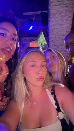 Group selfie of four friends at a lively nightclub bar with purple-blue neon lighting, one applying lip gloss, TVs and a textured stone wall in the background