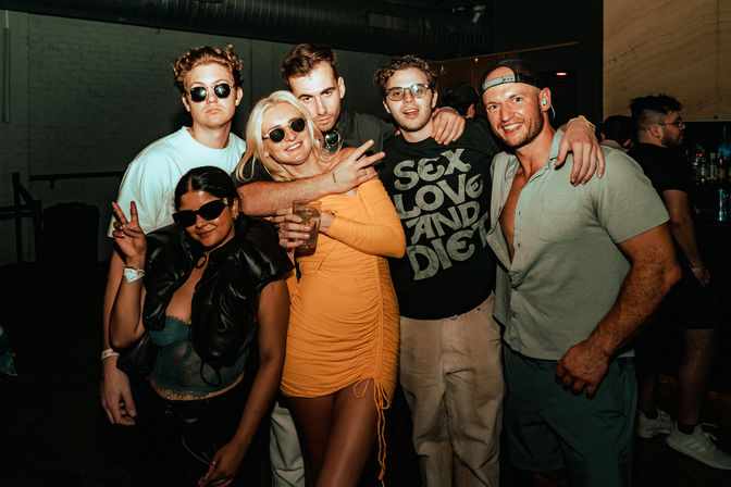 Group of six friends posing at an urban nightclub/bar with dim lighting — woman in a yellow dress holding a drink, others in casual clubwear and sunglasses, smiling and flashing peace signs.