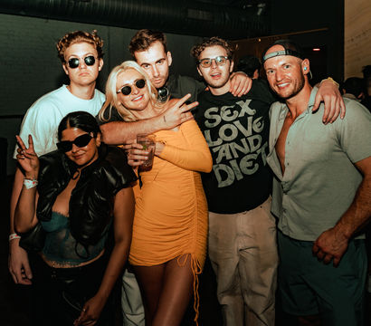 Group of six friends posing at an urban nightclub/bar with dim lighting — woman in a yellow dress holding a drink, others in casual clubwear and sunglasses, smiling and flashing peace signs.