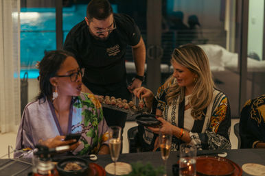 Sushi chef serving bite-sized rolls to two women at a lively evening dinner in a modern home dining room with an illuminated pool visible through glass.