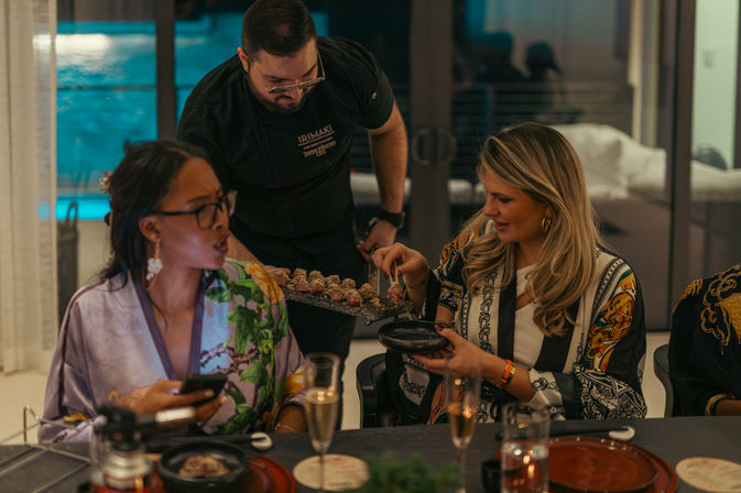 Sushi chef serving bite-sized rolls to two women at a lively evening dinner in a modern home dining room with an illuminated pool visible through glass.