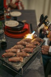 Culinary torch searing thin beef slices on a hot stone grill at a dimly lit dining table, flames caramelizing the meat beside plates and a glass.