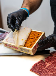 Gloved hands peeling paper from a wooden box of bright orange uni (sea urchin) next to marbled Wagyu beef slices on a white prep board, sushi-style prep.