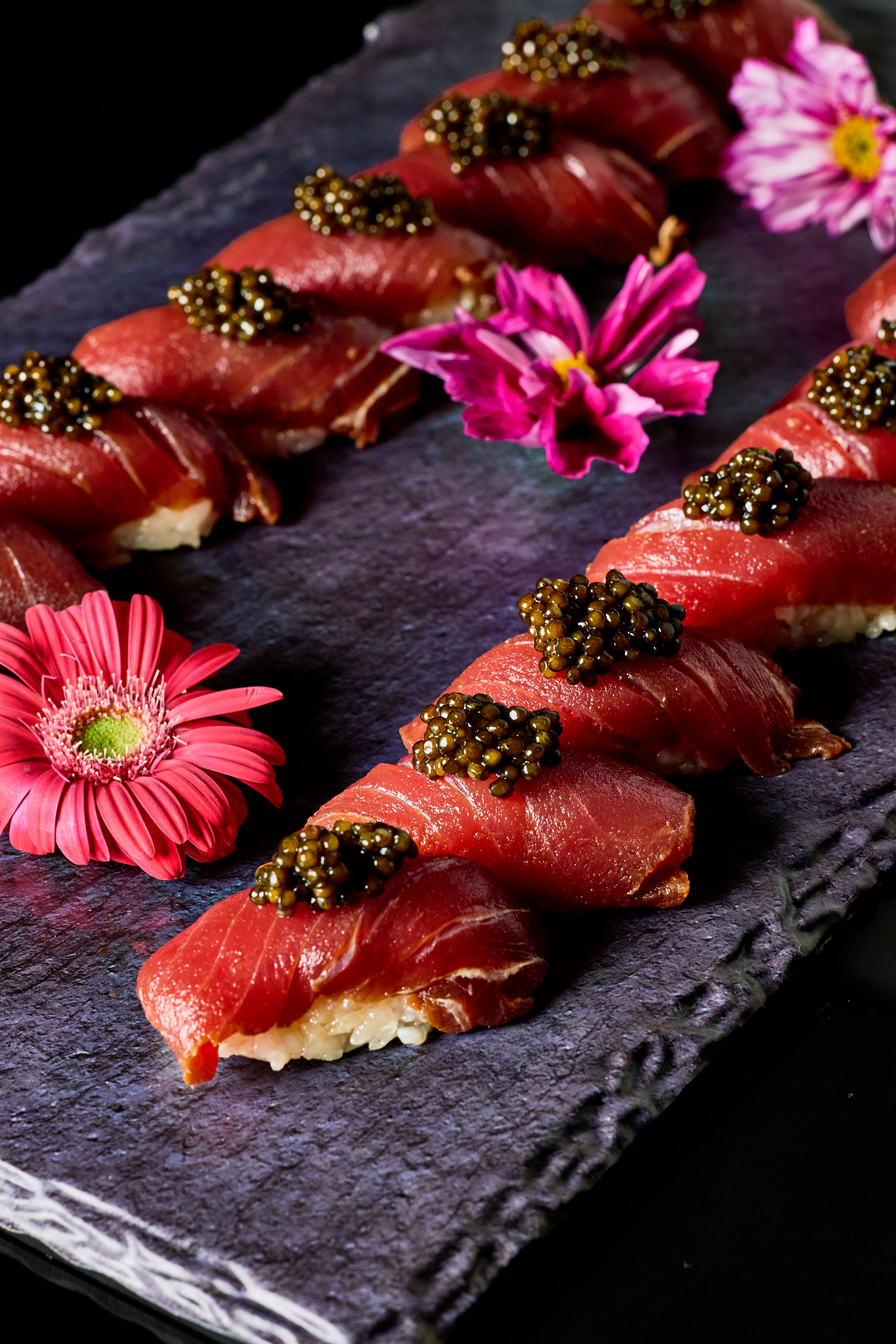 Close-up of gourmet tuna nigiri sushi topped with black caviar on a dark slate platter, accented by pink edible flowers — elegant Japanese-style plating.
