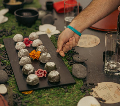 Hand reaching for powdered round dessert truffles on a black platter at an elegant dinner table with a moss runner, river stones, orchids, glassware and printed menus — zen-inspired centerpiece.