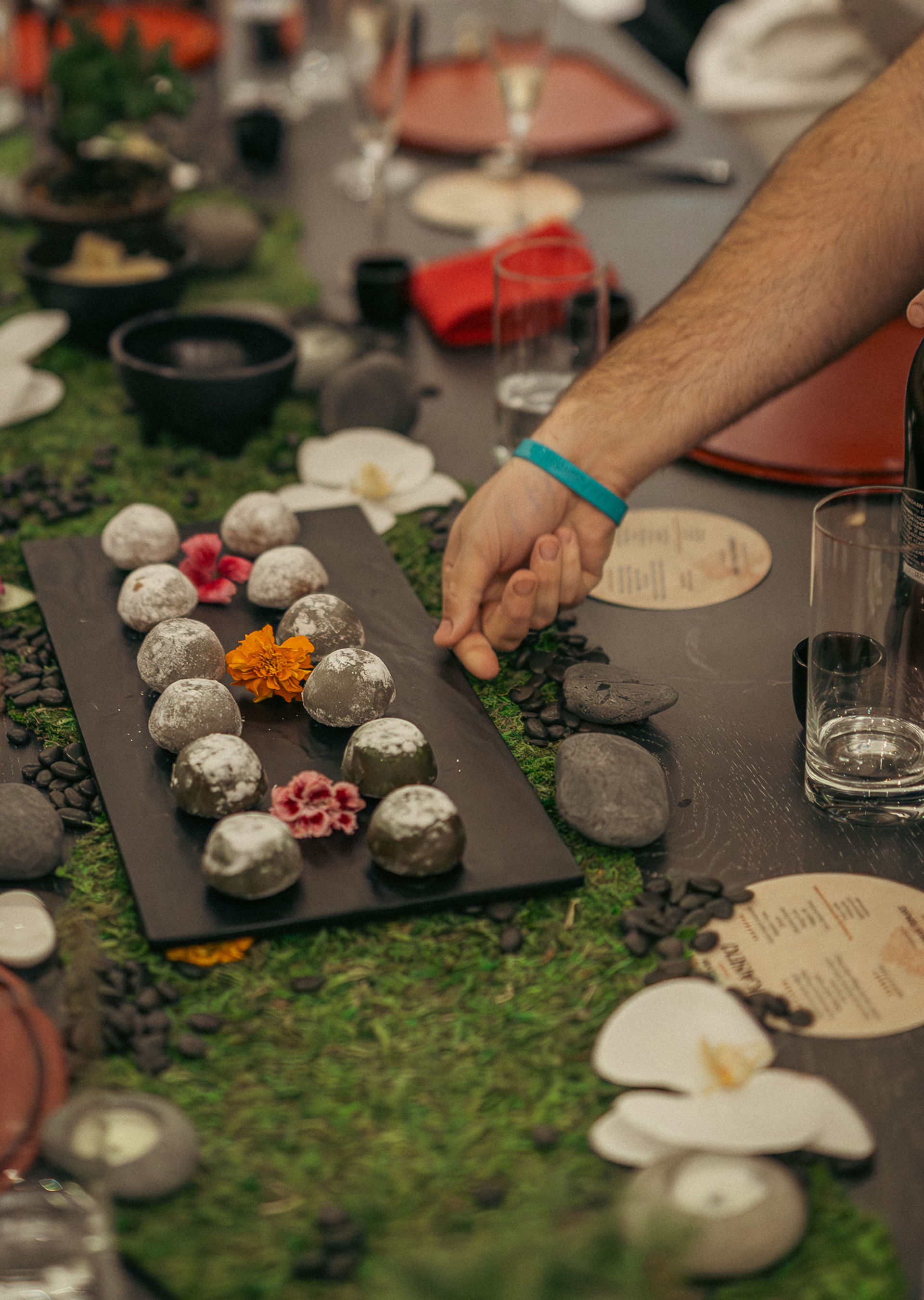 Hand reaching for powdered round dessert truffles on a black platter at an elegant dinner table with a moss runner, river stones, orchids, glassware and printed menus — zen-inspired centerpiece.